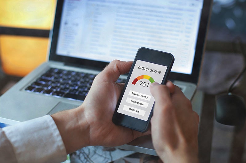 Hands holding a smartphone displaying a 751 credit score with a green gauge, in front of a laptop.