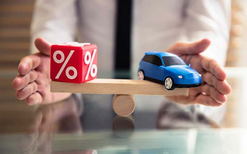 Hands balancing a red percent block and a blue toy car on a seesaw, symbolizing interest rates vs. car cost.